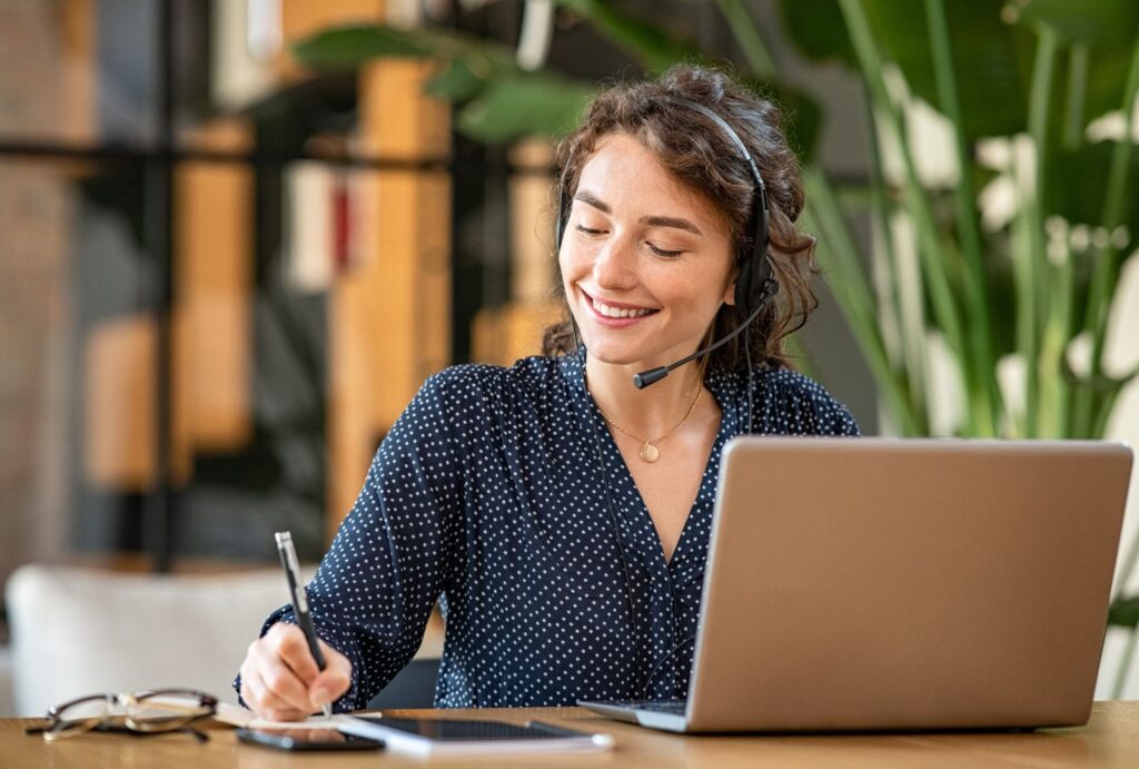 Kvinna med headset arbetar vid en laptop och antecknar under ett digitalt kundsamtal.